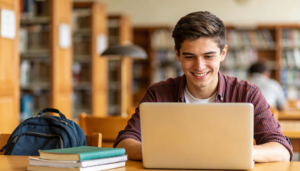 Ein lächelnder junger Mann sitzt an einem Bibliothekstisch mit einem Laptop, neben ihm ein Rucksack und gestapelte Notizbücher. Im Hintergrund sind mit Büchern gefüllte Regale zu sehen.