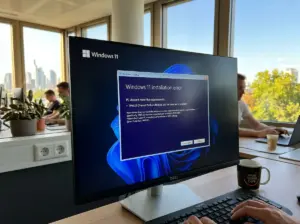 Auf einem Computerbildschirm wird eine Fehlermeldung zur Installation von Windows 11 angezeigt. Die Szene spielt in einem Büro, in dem Menschen an Schreibtischen arbeiten und durch große Fenster Bäume und Stadtgebäude zu sehen sind. Neben der Tastatur steht eine Kaffeetasse.